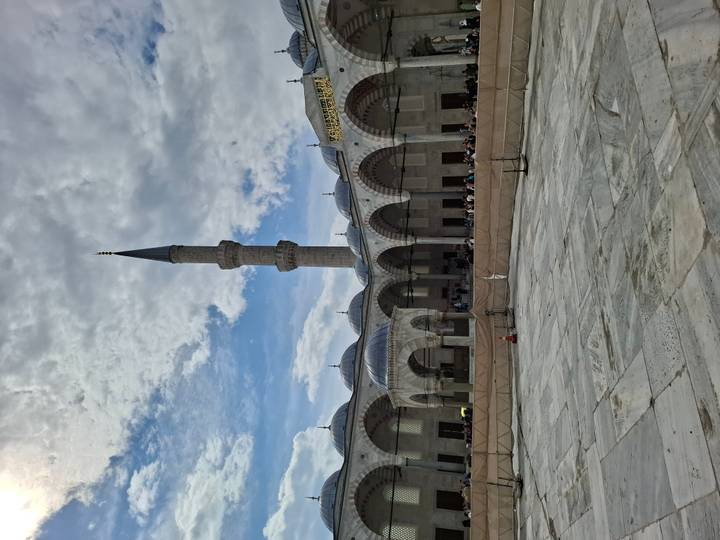 A grand mosque courtyard with elegant domes and a slender minaret against a partly cloudy sky.