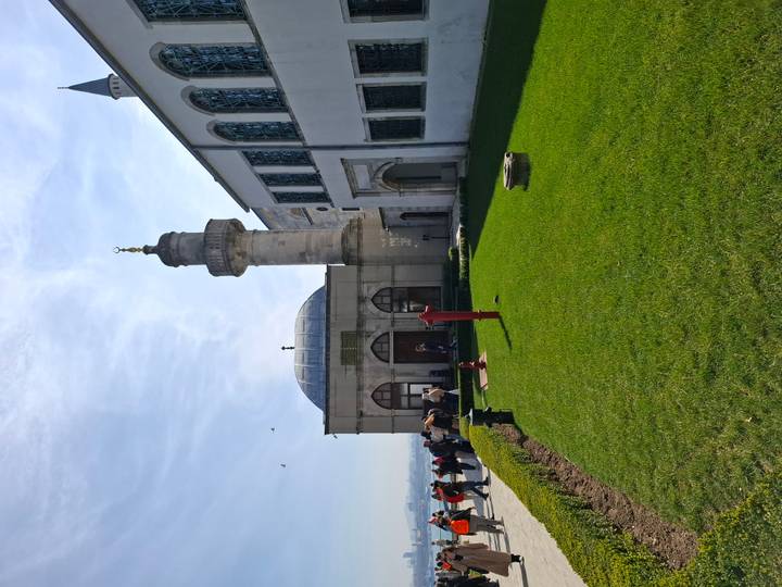 An exterior view of a small mosque with a green lawn and visitors milling about on a sunny day.