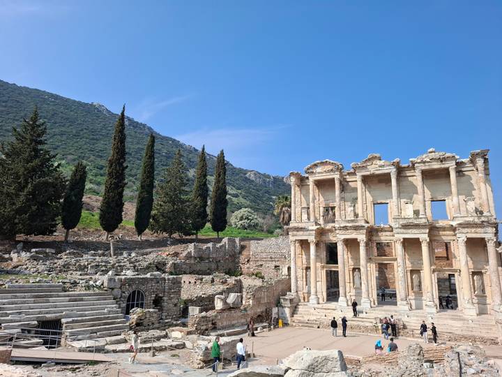 The well-preserved façade of the Library of Celsus stands beside tall cypress trees and ancient ruins.