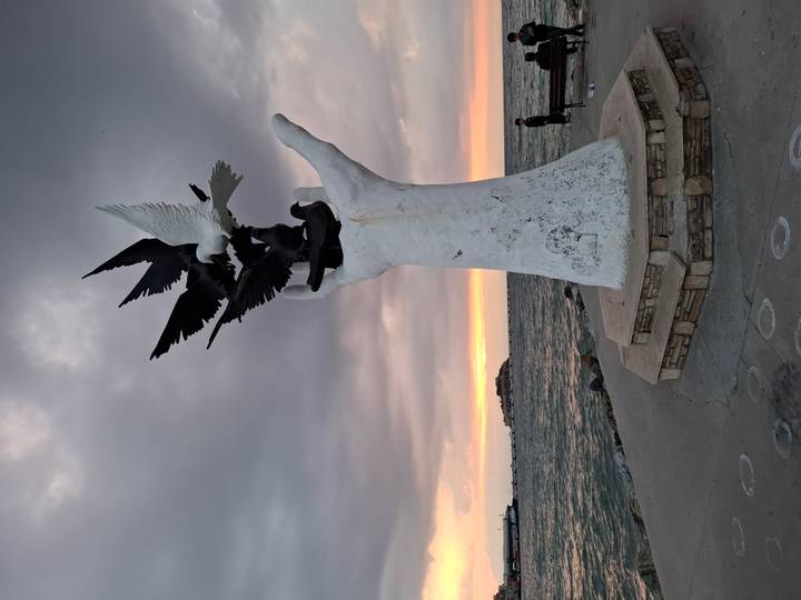 A sculptural hand holding birds overlooks the sea at dusk with an island in the distance.