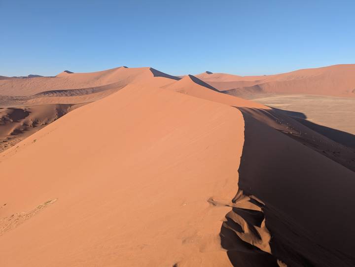 Sharp dune ridge casting long shadow across vast Namib Desert under crisp blue sky.