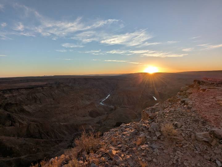 A dramatic view over a vast rocky canyon with a river far below, as the sun sets on the horizon under a clear sky.
