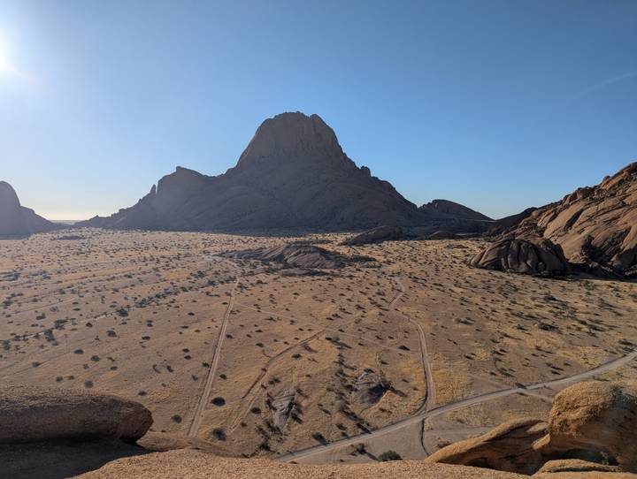 An isolated granite peak rises above an arid plain dotted with scrub under bright blue skies.