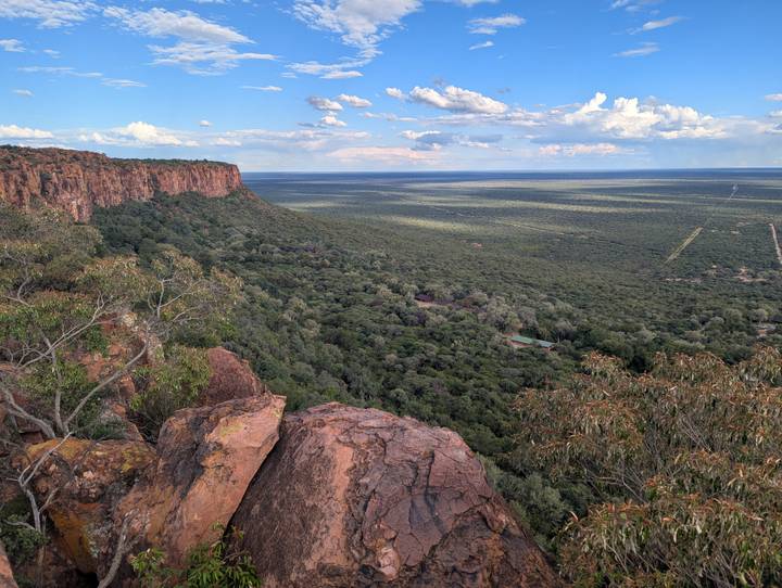 A lush green forest stretches to the horizon viewed from a rocky cliff edge under scattered clouds.