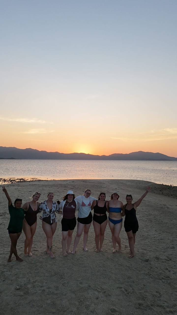 A group celebrates on a sandy spit as the sun sets behind distant mountains and calm sea.