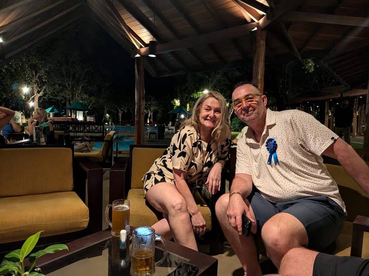 Smiling couple seated in an open-air lounge at night with a lit swimming pool behind.