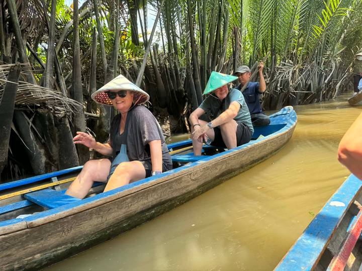 Tourists wearing conical hats paddle through narrow, palm-fringed canal in a wooden canoe