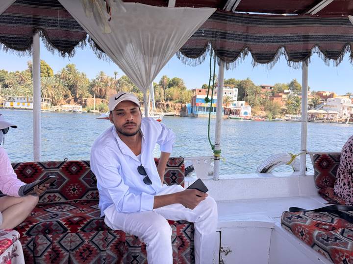 Man in white attire sits aboard a shaded felucca with colorful village on Nile bank behind