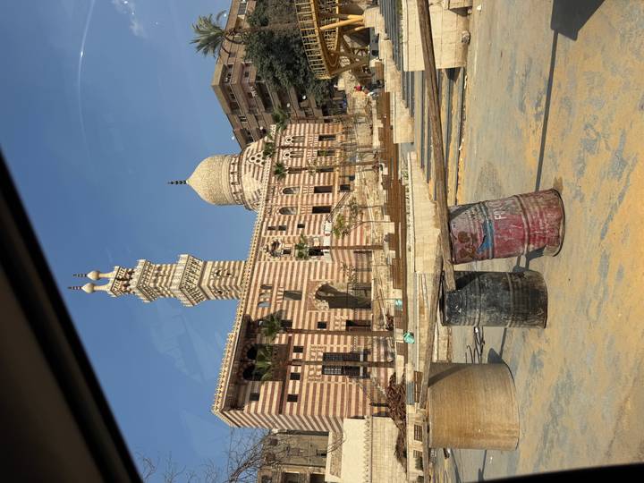Striped stone mosque with domes and minaret viewed beyond construction barrels