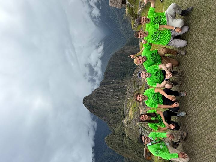 Six trekkers in green shirts posing at Machu Picchu with cloud-covered Huayna Picchu behind.