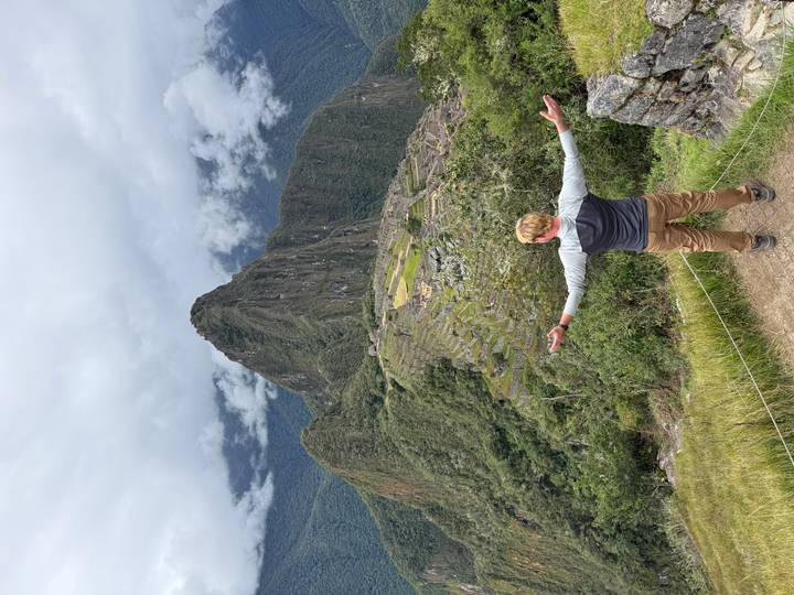 Solo traveller standing with arms outstretched overlooking Machu Picchu ruins on a cloudy day.