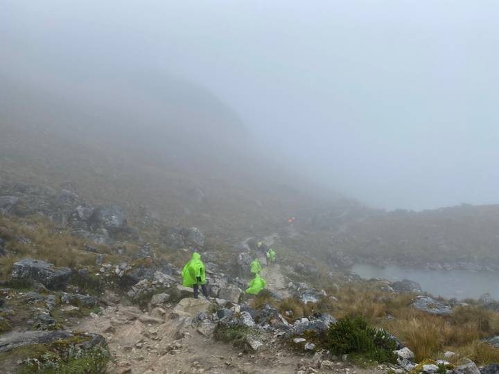 Hikers in bright ponchos walking through dense mountain fog on a rocky high-altitude trail.