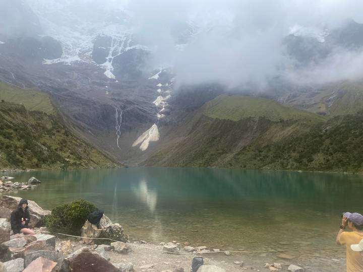 Turquoise glacial lake surrounded by steep green slopes and snow patches with trekkers resting at the shore.
