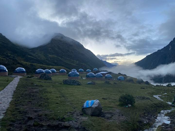 Row of blue-topped dome tents set on a grassy alpine valley with misty mountains at dawn.