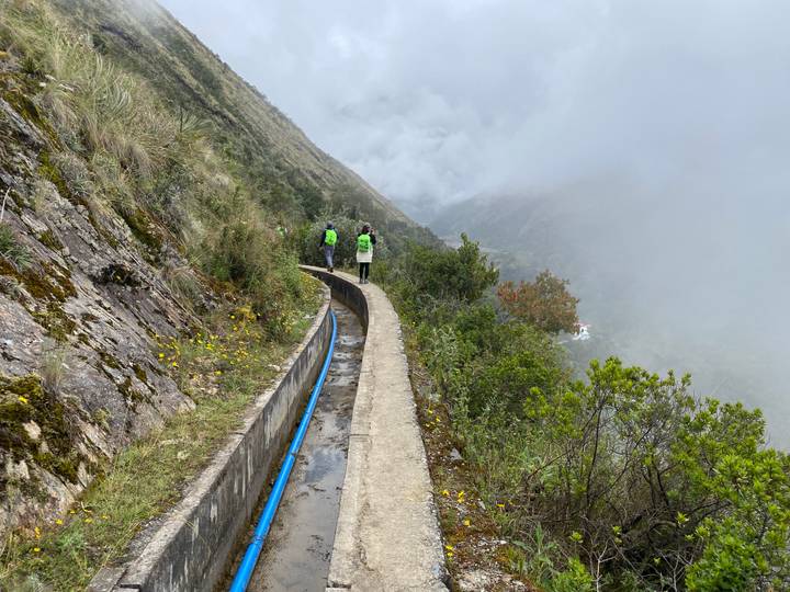 Trekkers in green vests walking along a narrow irrigation canal carved into a steep, mist-covered mountainside.