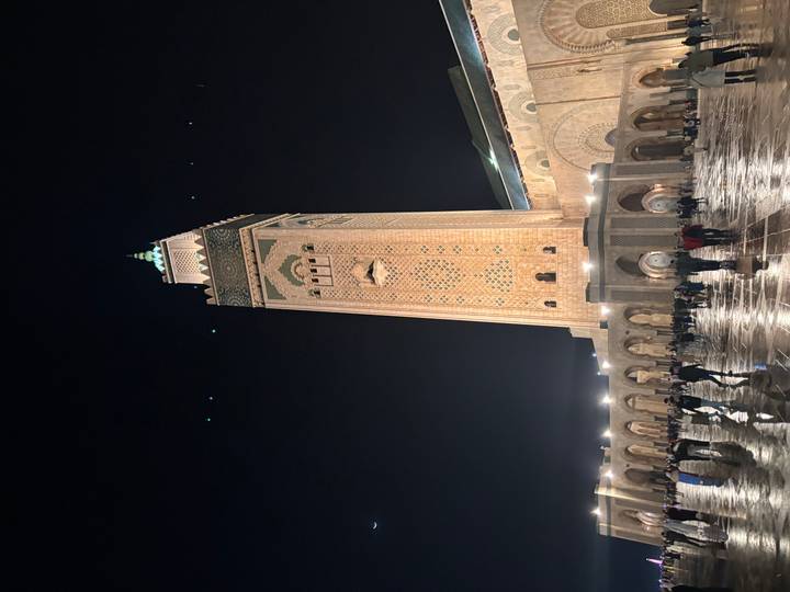 Night view of the illuminated Hassan II Mosque minaret towering over worshippers on the vast wet courtyard.