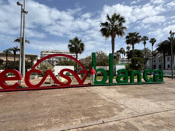 Traveler posing beside large red and green Casablanca sign with palms and blue sky overhead.