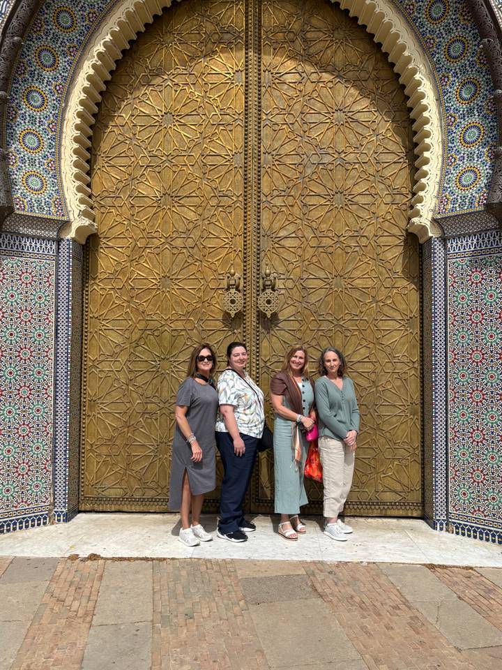Four women stand smiling in front of ornate golden doors of the Royal Palace in Fes.