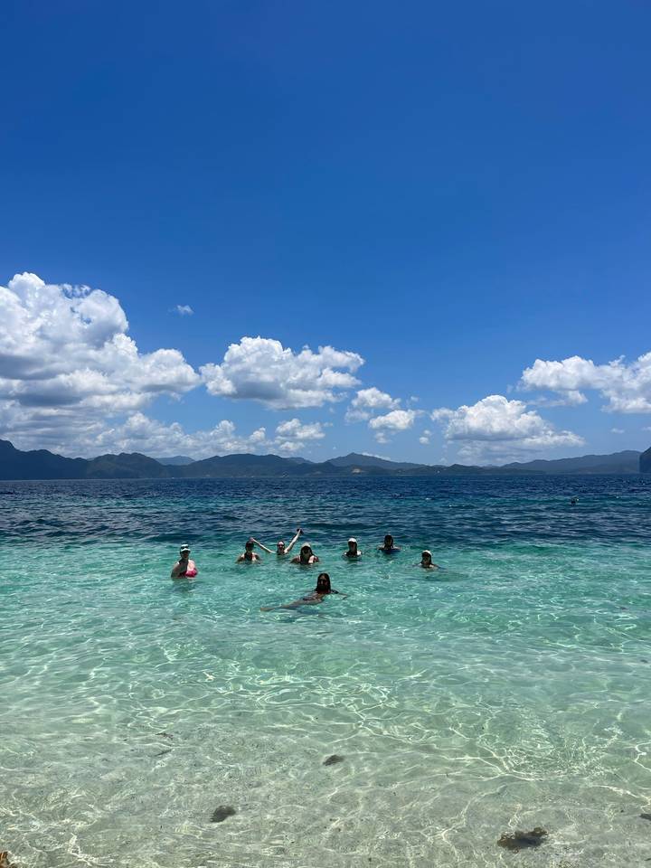 Group of travelers waist-deep in turquoise sea waving toward the camera with mountains on the horizon.