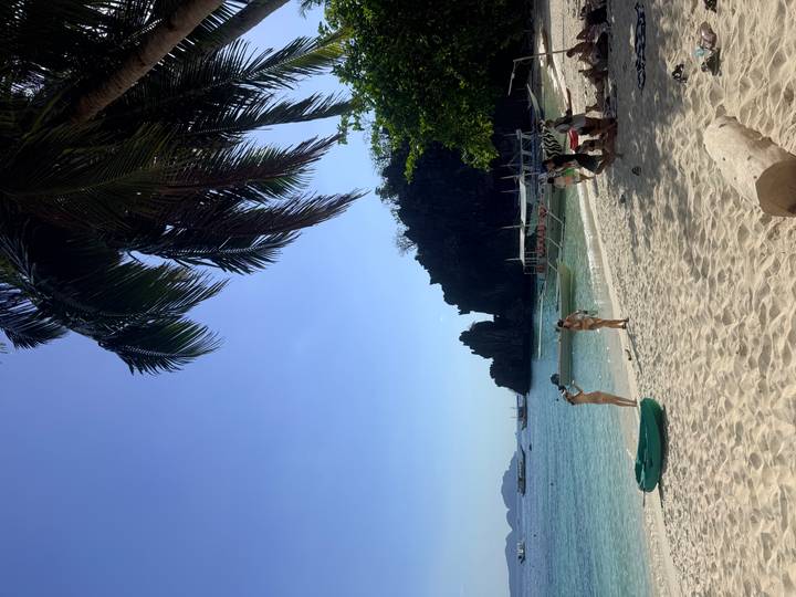 Tropical white-sand beach with turquoise water, limestone cliffs in the distance, small boats offshore and a few travellers beneath a leaning palm tree.