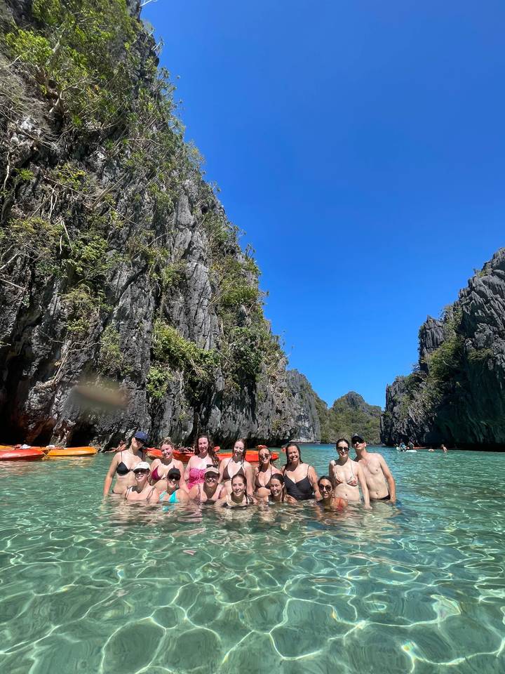 Group of friends posing waist-deep in a clear lagoon surrounded by towering limestone cliffs under a bright blue sky.