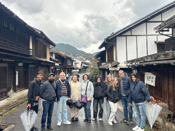 A tour group poses on a traditional wooden street flanked by dark-timber Edo-style houses with forested mountains in the background under a cloudy sky.