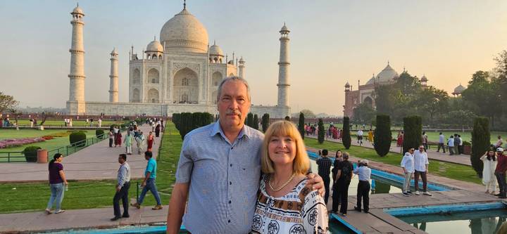 A smiling couple stands in the Taj Mahal gardens at dusk with the iconic mausoleum glowing softly behind.