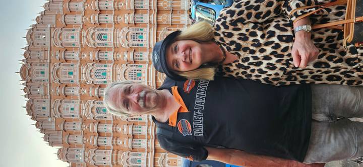 Couple stands before Jaipur’s ornate Hawa Mahal wearing casual attire and smiling at the camera.