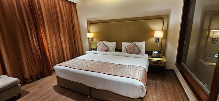Modern hotel room with king bed, warm lighting, brown wood floors and draped curtains.