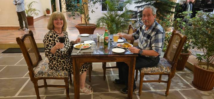 Couple enjoys dinner at a wooden table in a plant-filled restaurant, smiling toward the camera.
