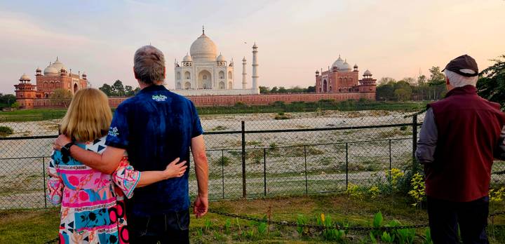 Travelers admire the Taj Mahal across the river at sunset, standing by a fence with warm sky tones.