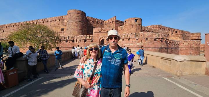 Couple poses on the bridge leading into the massive red-sandstone walls of Agra Fort under a clear blue sky.