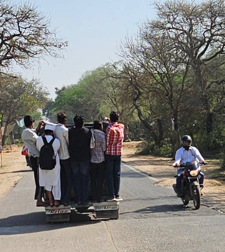 Blurry shot of several men riding on the back of a vehicle along a rural tree-lined road.