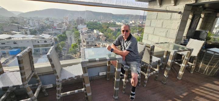 Traveler relaxes at a rooftop glass-panel bar overlooking a sprawling city and surrounding hills.