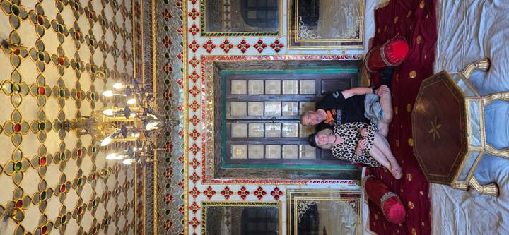 Couple seated on rich red cushions in an opulent palace room decorated with mirrors and chandeliers.