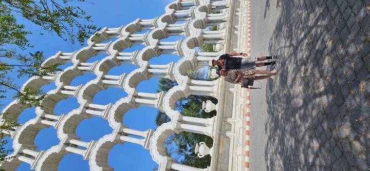 Travelers stand beneath a towering white multi-arch installation against a vivid blue sky.