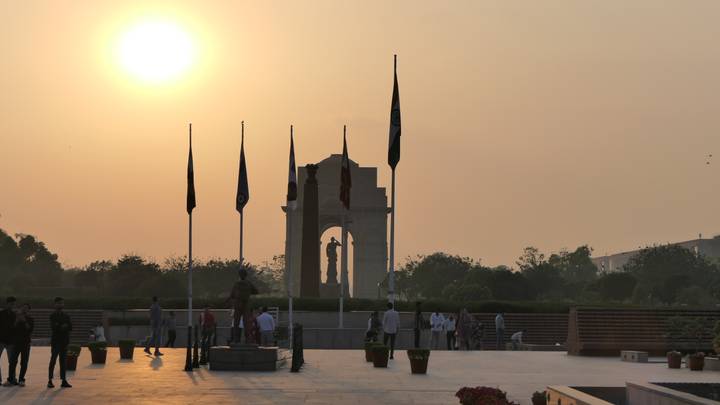 Silhouetted India Gate memorial and flagpoles at dusk with a glowing sun low in the hazy sky.