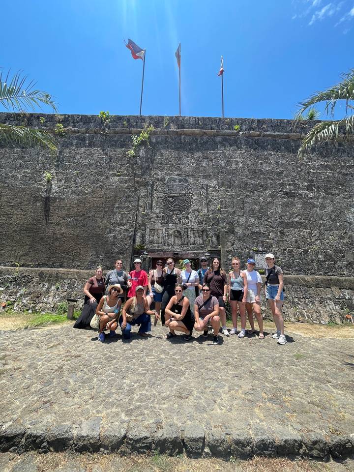 Tour group poses in front of a massive stone fort wall under harsh midday sun.