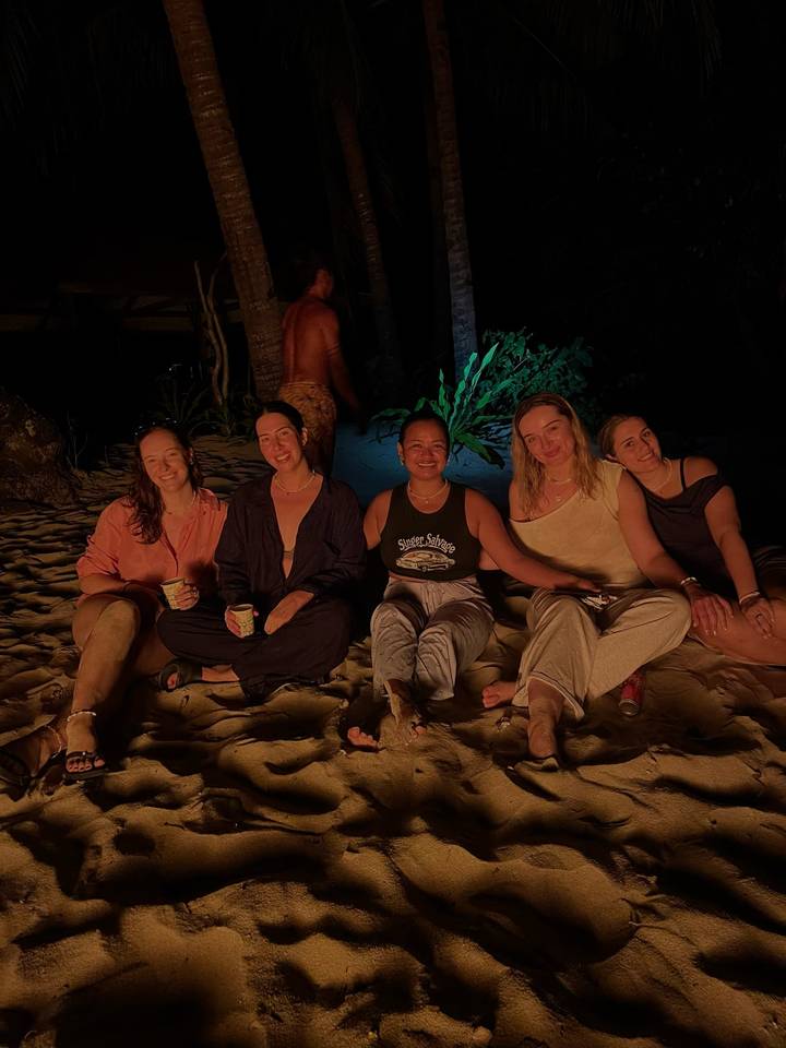 Group of friends sit on a beach around a fire, warmly lit against the dark night background.