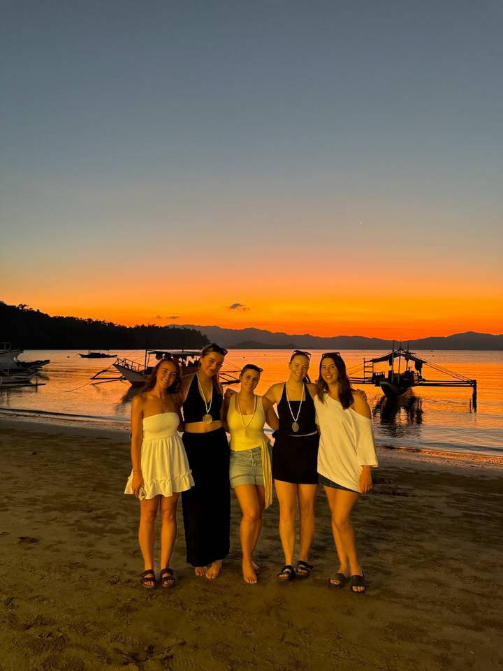 Five women pose on the beach at vivid orange sunset with boats and mountains in the distance.