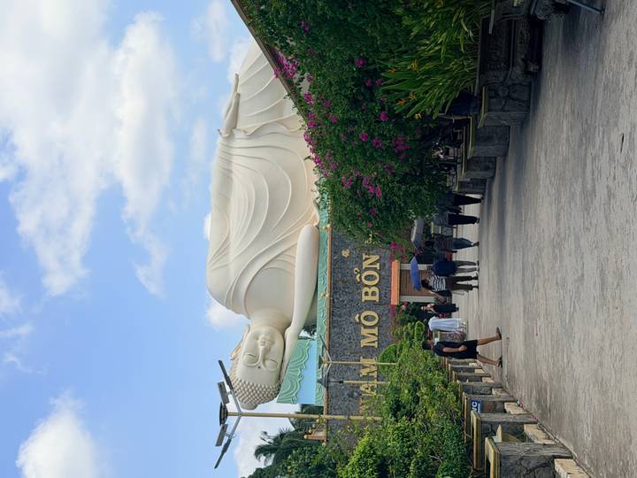Large reclining Buddha statue towers above visitors walking through a bougainvillea-lined courtyard.