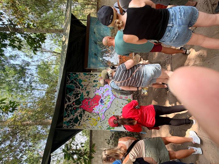Visitors listen to a guide beside a large illustrative map display at an outdoor museum, finger partially visible.