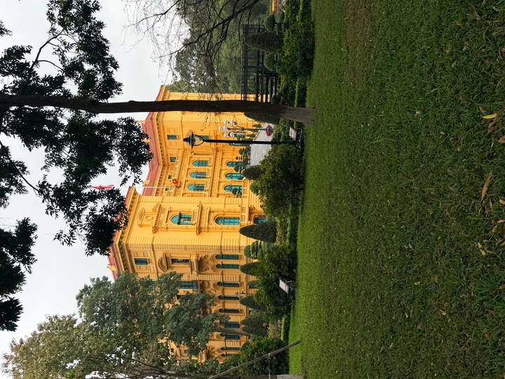 Bright yellow French-colonial palace surrounded by manicured lawns and tall shade trees in a city park.
