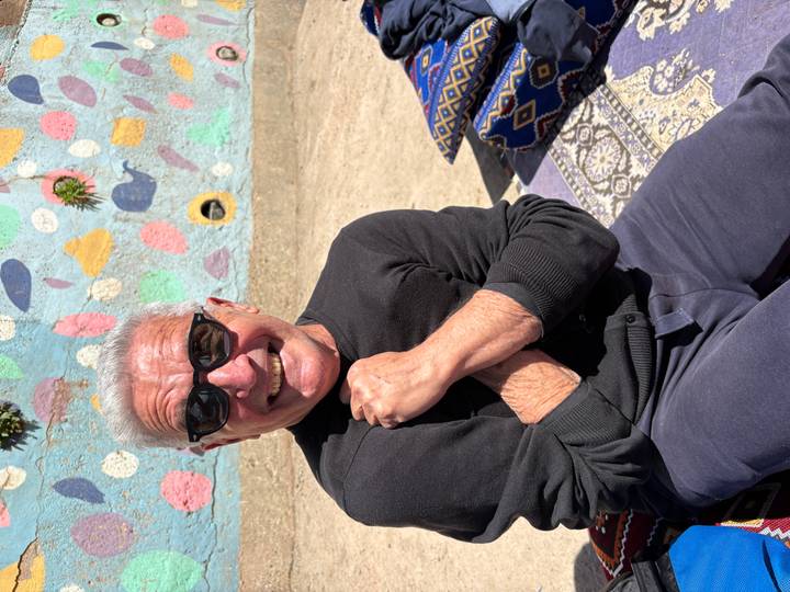 Close portrait of a smiling man wearing sunglasses seated against a colorful mural wall.