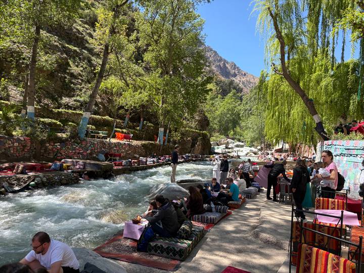 Bustling outdoor cafe area lining a rushing river under leafy trees and mountain cliffs.