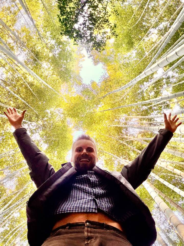 A smiling traveler stretches arms upward beneath towering bamboo stalks.