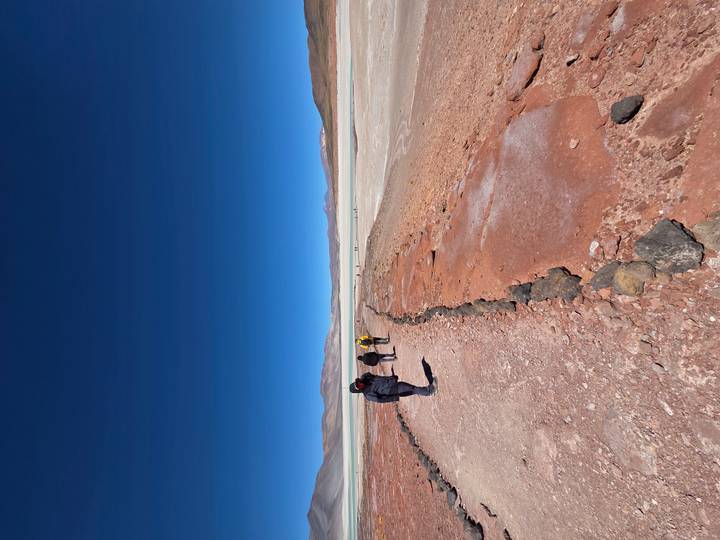 Hikers walk along a red-earth trail toward a wide white salt flat under a deep blue sky.