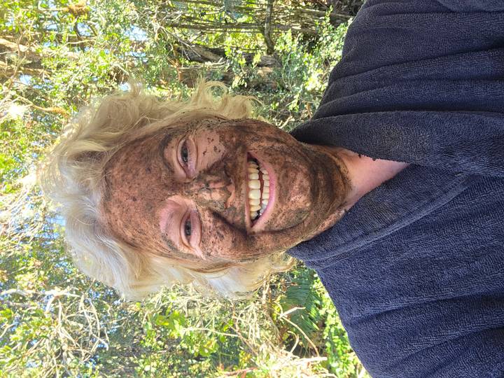 A smiling woman covered in a playful mud mask stands outdoors in a robe.