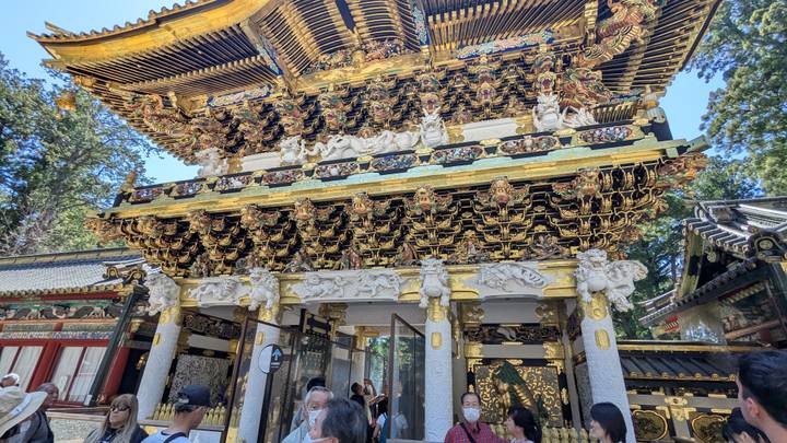 Crowds gather before an ornate gold-covered temple gate surrounded by tall trees.