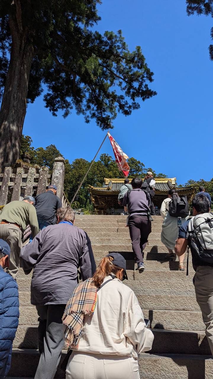Visitors ascending stone steps toward a shrine roof while a guide holds a flag under a clear blue sky.